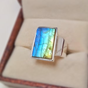 May include: A silver ring with a large, rectangular, iridescent labradorite gemstone. The gemstone has shades of blue, green, and yellow. The ring is set in a simple, modern style.