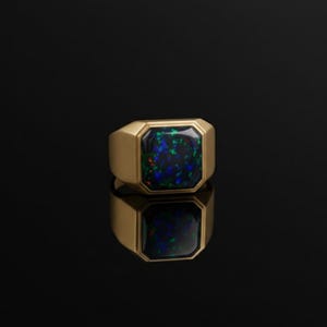 May include: A gold ring featuring a rectangular black opal gemstone. The opal displays blue and green flecks. The ring is set on a reflective black surface.