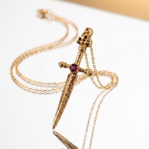 Può includere: Una collana color oro con un ciondolo a forma di piccola spada o pugnale. L'elsa della spada è decorata con una singola pietra preziosa rotonda di colore rosso intenso al centro della guardia. La superficie metallica presenta una finitura ruvida.
