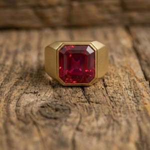 Puede incluir: Un anillo dorado con una gran gema roja rectangular facetada. El anillo tiene una banda ancha y un acabado de metal cepillado. La gema está engastada en un bisel, creando una pieza audaz y elegante.