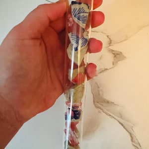 May include: A clear glass vial filled with small, colorful fragments of what appears to be pottery or ceramic. The fragments include blue and white patterned pieces, along with red and beige elements. The vial is sealed with a cork stopper.