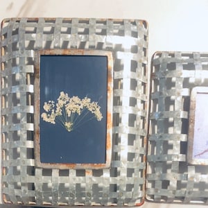 May include: Two rustic metal picture frames with a wire mesh design. The left frame has a pressed flower inside, while the right frame has a purple and blue abstract design.