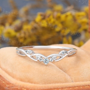 May include: A delicate, curved silver ring with a series of small, clear gemstones. The ring features an intricate design with interwoven patterns and a central cluster of stones. The ring is set against a soft, peach-colored background.