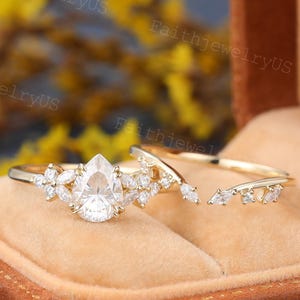 Könnte beinhalten: Ein goldfarbenes Ring-Set mit einem birnenförmigen Diamanten als Mittelstein, akzentuiert durch kleinere Diamanten. Das Band ist mit zusätzlichen Diamantclustern verziert. Das Set enthält ein passendes, gebogenes Band, alles in einer braunen Ringschatulle präsentiert.