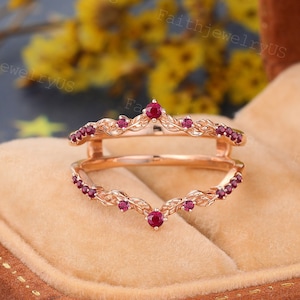 以下が含まれることがあります： V字型のデザインのローズゴールドのリングガード。リングガードには、小さくて丸い赤い宝石が飾られています。リングガードは茶色のジュエリーボックスにセットされています。