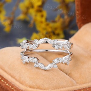 May include: A delicate, white gold ring enhancer with a curved, v-shaped design. The enhancer is adorned with small, round and marquise-shaped diamonds, creating a sparkling and elegant look.