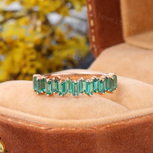 Pode incluir: Um anel de banda de ouro rosa com uma fileira de pedras preciosas de cor verde esmeralda em forma de baguette. O anel é engastado em forma curva e é apresentado em uma caixa de veludo marrom.