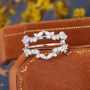 May include: A delicate silver ring enhancer with a marquise and round diamond design. The enhancer is set on a brown velvet background.
