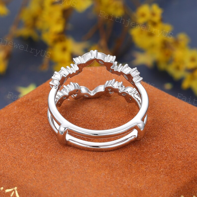 May include: A silver double ring with a delicate, curved design featuring small, clear stones. The ring is on a brown, textured surface.