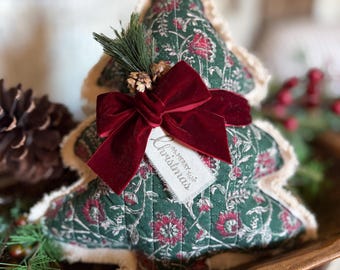 May include: A quilted fabric Christmas tree ornament in dark green with a red and white floral pattern. It features a frayed cream-colored edge, a deep red velvet bow, a small pine sprig, and a tag with the text Merry Christmas.