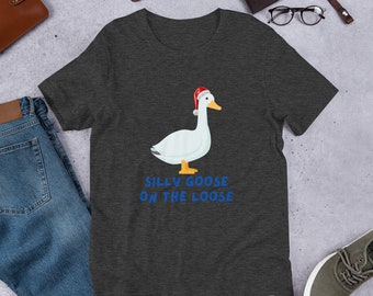 May include: A dark heather grey t-shirt with a white goose wearing a red Santa hat. The text "Silly Goose On The Loose" is printed in blue below the goose.