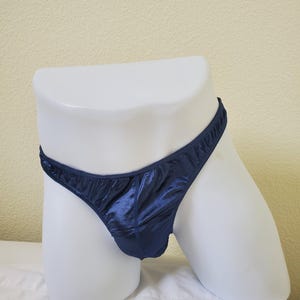 Könnte beinhalten: Ein dunkelblauer String-Tanga aus glänzendem, seidigem Material. Die Unterwäsche wird auf einer weißen Schaufensterpuppe präsentiert. Der Stoff hat einen leichten Glanz, und die Kanten sind sauber verarbeitet.