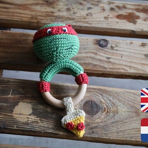 Op de afbeelding: Groene gehaakte babyratel met een rood bandana, houten ring en een gehaakte pizza-slice charme.