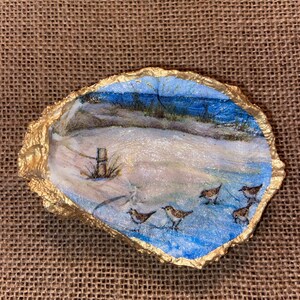 May include: A hand-painted oyster shell with a blue and white seascape scene. The scene includes a sandy beach, a wooden fence post, and a flock of small brown birds walking on the sand. The shell is trimmed with gold paint.