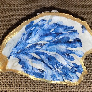 May include: A white oyster shell with a blue and white painted design of seaweed. The shell is accented with a gold rim.