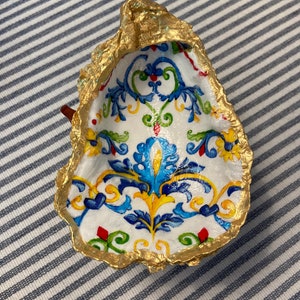 Op de afbeelding: Een witte keramische schaal met een gouden rand en een blauw, geel en groen bloemenpatroon. De schaal is schelpvormig.