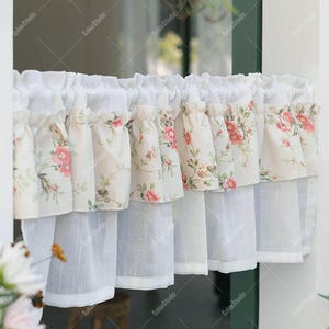 Puede incluir: Cortina de ventana blanca con estampado floral. La cortina presenta un diseño en capas con una parte superior con volantes y un estampado floral en tonos rosa y verde. La capa inferior es de tela blanca transparente.