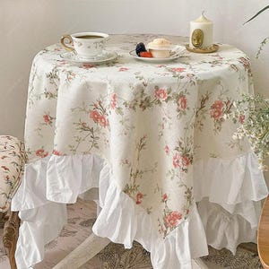 May include: A round table covered with a cream-coloured tablecloth featuring a pink and green floral pattern and a white ruffled hem. On the table sits a white teacup on a saucer, a small plate with a dessert, and a white pillar candle.
