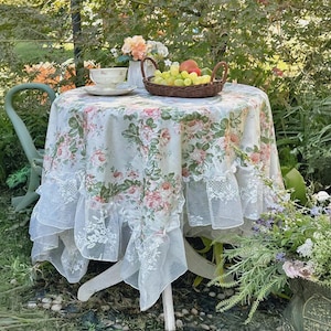 May include: A round table with a floral tablecloth, adorned with pink roses and green foliage, and a lace trim. A wicker basket of fruit and a teacup and saucer are on the table. A green chair is positioned nearby.