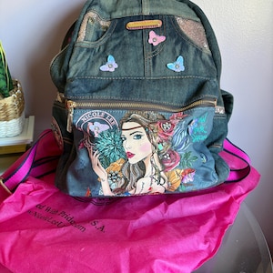 May include: Denim backpack with a colorful illustration of a woman with flowers and butterflies. The backpack has a front zippered pocket, pink butterfly accents, and a pink and black strap. A pink dust bag is included.