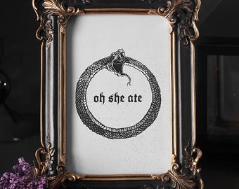May include: A decorative picture frame in black and gold, displaying a white print with an ouroboros illustration and the words "oh she ate." The frame has intricate detailing and is set against a dark backdrop, with purple flowers.