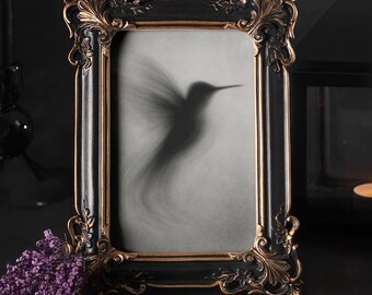 May include: Ornate black picture frame with gold accents, featuring a hummingbird silhouette. The frame has detailed scrollwork and a rectangular shape. A sprig of purple flowers rests near the frame, with a dark background.