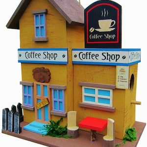 Könnte beinhalten: Ein Vogelhaus aus Holz in Form eines Cafés mit einem roten Dach, blauen Fenstern und einem Schild mit der Aufschrift "Coffee Shop". Vor dem Laden stehen ein kleiner Tisch und zwei Stühle.