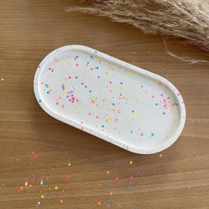 Könnte beinhalten: Ovale, weißes Tablett mit bunten, konfettiartigen Tupfen. Das Tablett hat abgerundete Kanten und besteht aus einem festen Material. Das Konfetti umfasst Pink, Orange, Gelb und Blau. Das Tablett liegt auf einer Holzoberfläche.