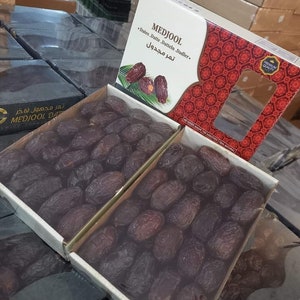 May include: Two boxes filled with Medjool dates. The boxes are labelled "Medjool Dates" and "تمر مجدول". The dates are a deep brown colour and are arranged in a neat pattern.