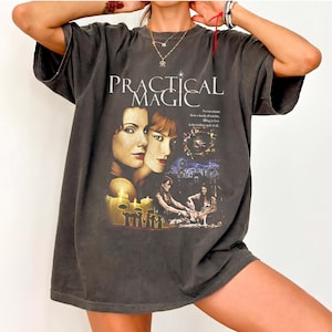 May include: A charcoal gray short-sleeve t-shirt featuring a graphic print with the text "PRACTICAL MAGIC" above portraits of two individuals, a small circular image, and a scene of two figures with candles, all in a vintage-inspired style.