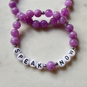 May include: Two beaded bracelets. The bracelets are made of round, light purple beads and silver-colored spacers. One bracelet spells out "SPEAK NOW" with white letter beads. The bracelets are on a white marble surface.