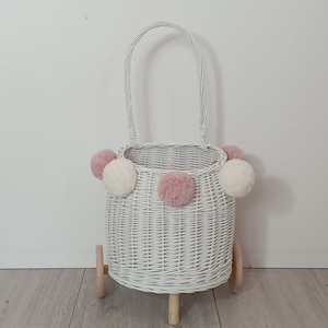 Puede incluir: Cesta de mimbre blanca con ruedas y asa. La cesta está decorada con pompones rosas y blancos.