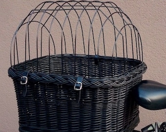 Op de afbeelding: Zwarte rieten fietsmand met een zwarte metalen kooi bovenop. De mand heeft twee leren riemen met gespen aan de voorkant.
