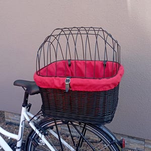Puede incluir: Una cesta de bicicleta de mimbre negra con un forro de tela rojo y una parte superior de jaula de metal negra. La cesta está unida a la parte trasera de una bicicleta blanca.