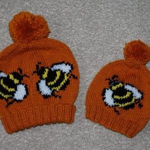 Peut inclure: Deux bonnets en tricot orange avec des pompons et des motifs d'abeilles. Le plus grand bonnet a deux abeilles, tandis que le plus petit bonnet en a une.