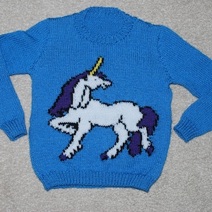 Puede incluir: Un suéter de punto azul con un diseño de unicornio blanco. El unicornio tiene una melena y una cola moradas y un cuerno dorado.