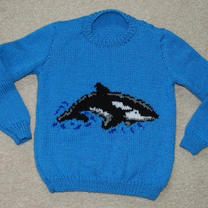 Puede incluir: Un suéter azul tejido con un diseño de orca en blanco y negro. La orca se representa nadando en olas azules. El suéter tiene mangas largas y cuello redondo.