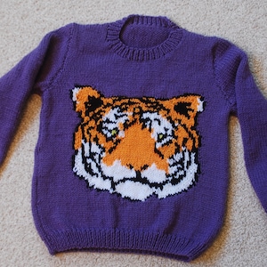 Op de afbeelding: Paarse gebreide trui met een tijgergezichtontwerp in oranje, wit en zwart.