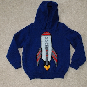Könnte beinhalten: Blauer gestrickter Kapuzenpullover mit einem Raketenmotiv in Weiß, Rot und Grau. Die Rakete hat eine gelbe Flamme.