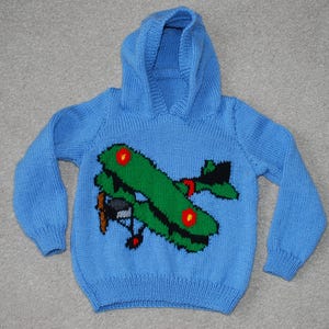 May include: A light blue knitted hoodie featuring a green airplane design with red and black accents. The hooded sweater is a children's garment, perfect for casual wear. The airplane has a propeller and is centered on the front.