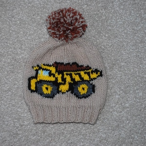 May include: A beige knitted hat with a brown and white pom pom on top. The hat has a yellow and black knitted design of a dump truck on the front.
