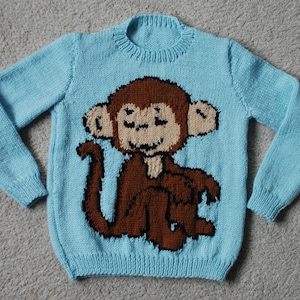 Könnte beinhalten: Hellblauer Strickpullover mit einem braunen Affenmuster auf der Vorderseite. Der Affe hat ein lächelndes Gesicht. Der Pullover hat lange Ärmel und einen gerippten Ausschnitt und Bündchen. Das Kleidungsstück ist aus Strickstoff.