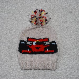 Op de afbeelding: Een beige gebreide muts met een rood en zwart raceauto-ontwerp en een kleurrijke pompon bovenop.
