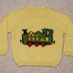 Könnte beinhalten: Ein gelber Strickpullover mit einem grünen, roten und schwarzen Zugmotiv auf der Vorderseite.