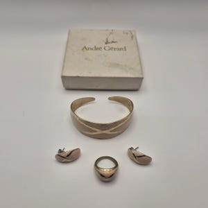 May include: A jewellery set including a cuff bracelet, a ring, and two stud earrings, all featuring a light pink enamel finish with gold-tone accents. The items are displayed with a square white box labelled with the text André Gérard.