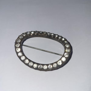 May include: Oval brooch with a silver-tone metal frame. The frame is set with numerous clear, sparkling rhinestones. A straight pin is attached to the back for securing the brooch to clothing or accessories.