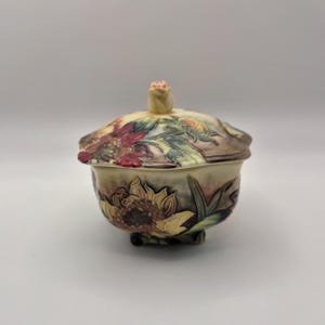 May include: A ceramic sugar bowl with a lid featuring a sculpted rose finial. The bowl and lid are decorated with embossed, hand-painted sunflowers in shades of yellow, deep red, and green against a cream-coloured background with dark shading.