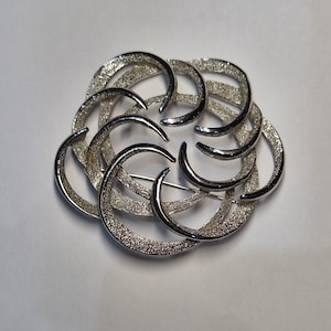 Op de afbeelding: Een zilverkleurige broche met een cirkelvormige opstelling van overlappende, halvemaanvormige segmenten. Elk segment heeft een getextureerd, korrelig oppervlak met gladde, gepolijste randen, wat een gelaagd, wervelend patroon in een ronde vorm creëert.
