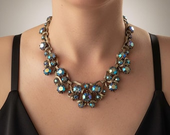 May include: A gold-toned metal necklace featuring a series of round, iridescent blue and teal faceted stones. The stones are set in a decorative, curved metal frame that forms a collar-style design, with a larger cluster of stones at the center.