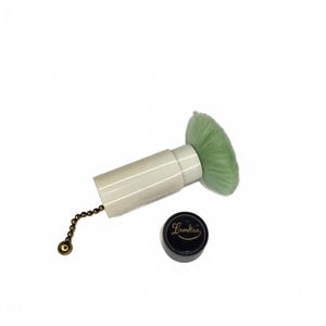 Può includere: Un pennello per il trucco con una testina verde chiaro e un manico bianco. Un piccolo contenitore nero con la scritta "Lamkin" in lettere dorate è accanto al pennello. Una catena dorata è attaccata al manico del pennello.